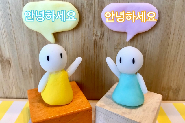 黄色と水色のワンピースを着た白いキャラクターが木の台に並んで立ち、吹き出しで韓国語の会話をしている様子のイラスト